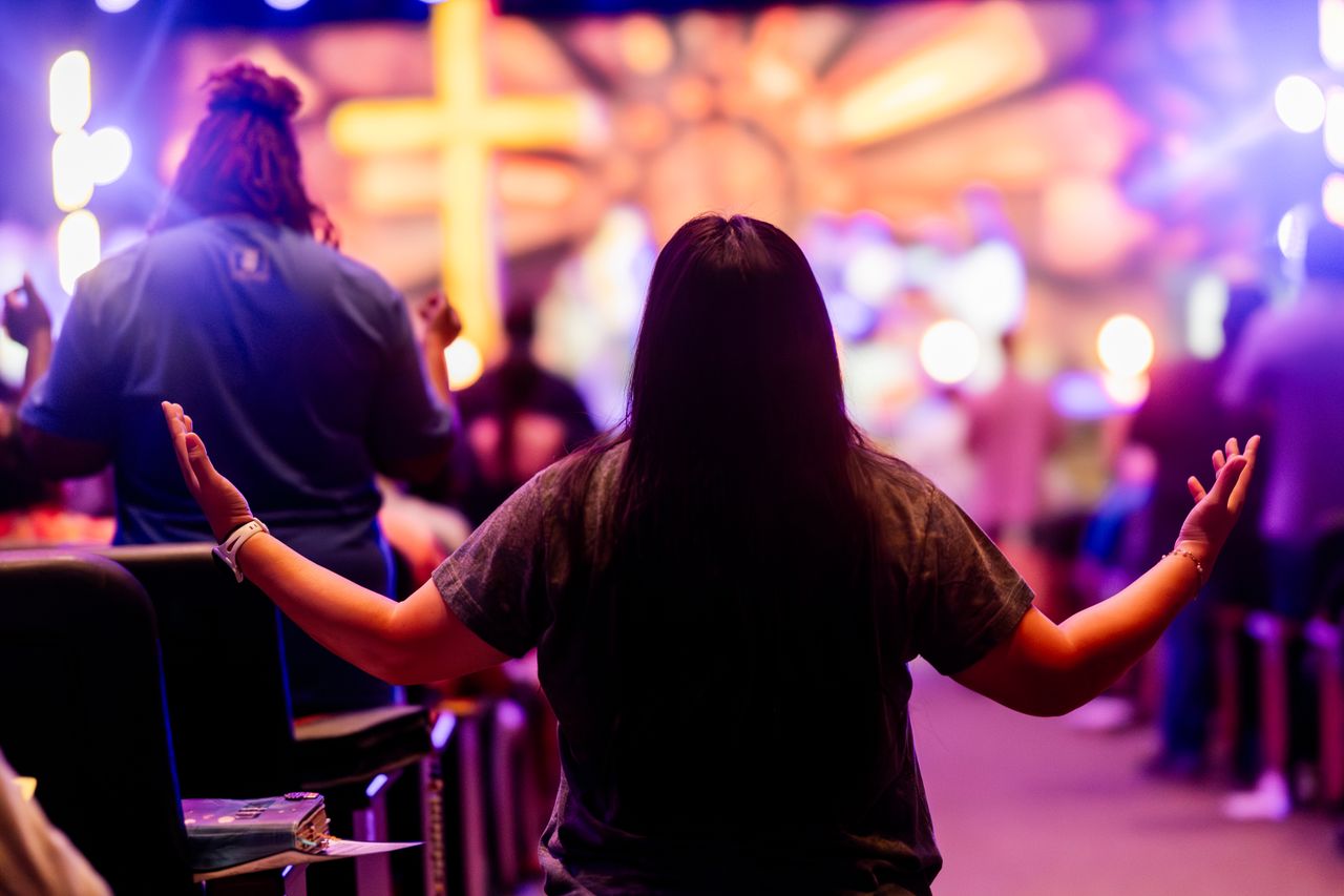 About Back view of people engaged in a vibrant religious service with colorful lighting and a cross.
