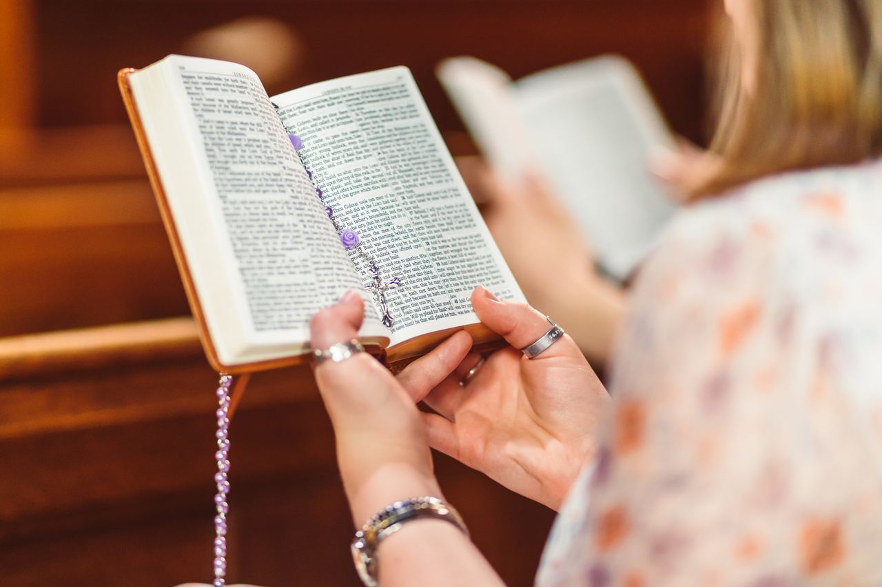 About A woman holding a Bible and rosary beads, engaged in prayer inside a church.